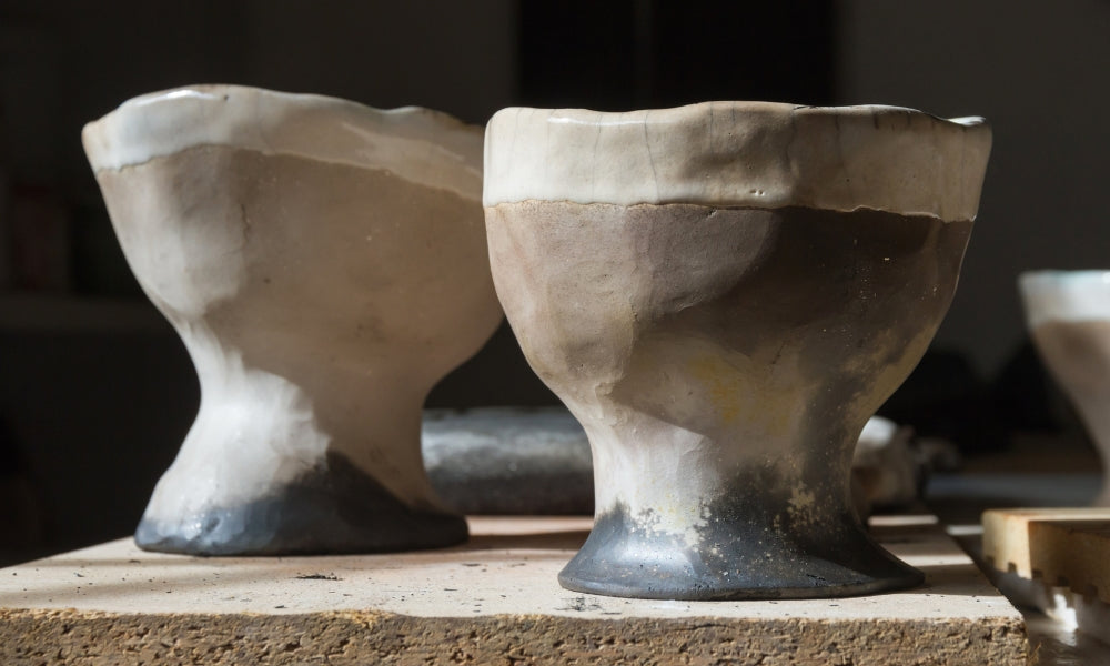 Dos cuencos de té de cerámica raku (chawan) con formas modeladas a mano y tonos de esmalte de oscuro a claro, sobre una superficie de taller