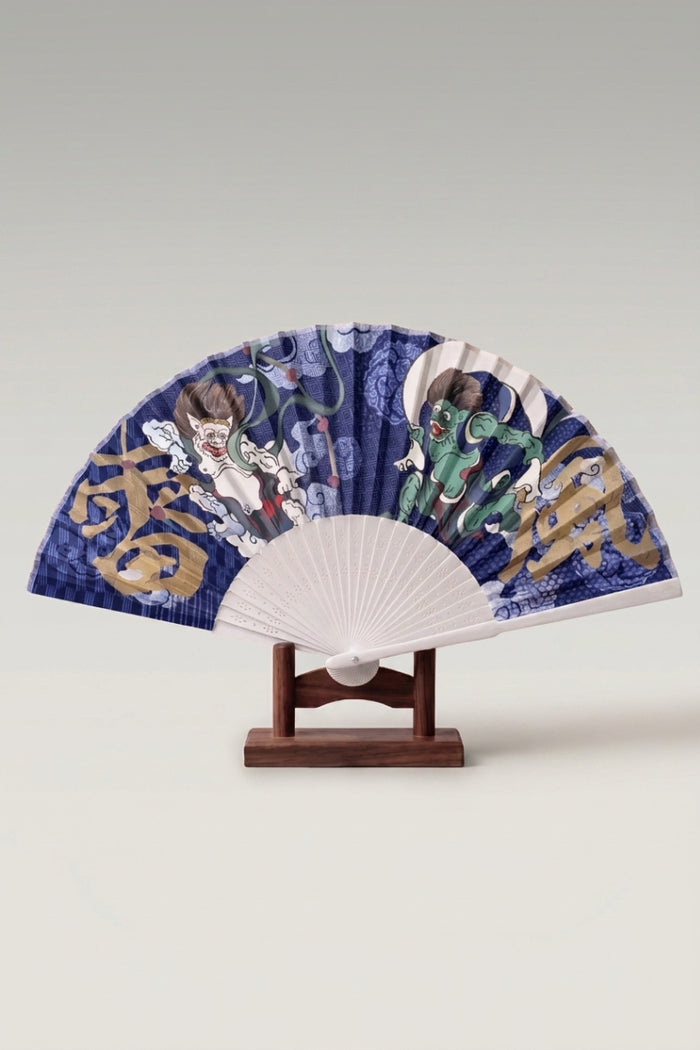 Abanico plegable japonés 'Fuurai' abierto sobre su soporte — Fujin y Raijin a ambos lados de caracteres dorados sobre hoja índigo
