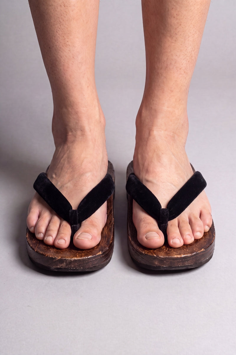 Chanclas japonesas color negro liso, calzadas descalzas de frente — suela de madera maciza marrón y tira acolchada entre los dedos