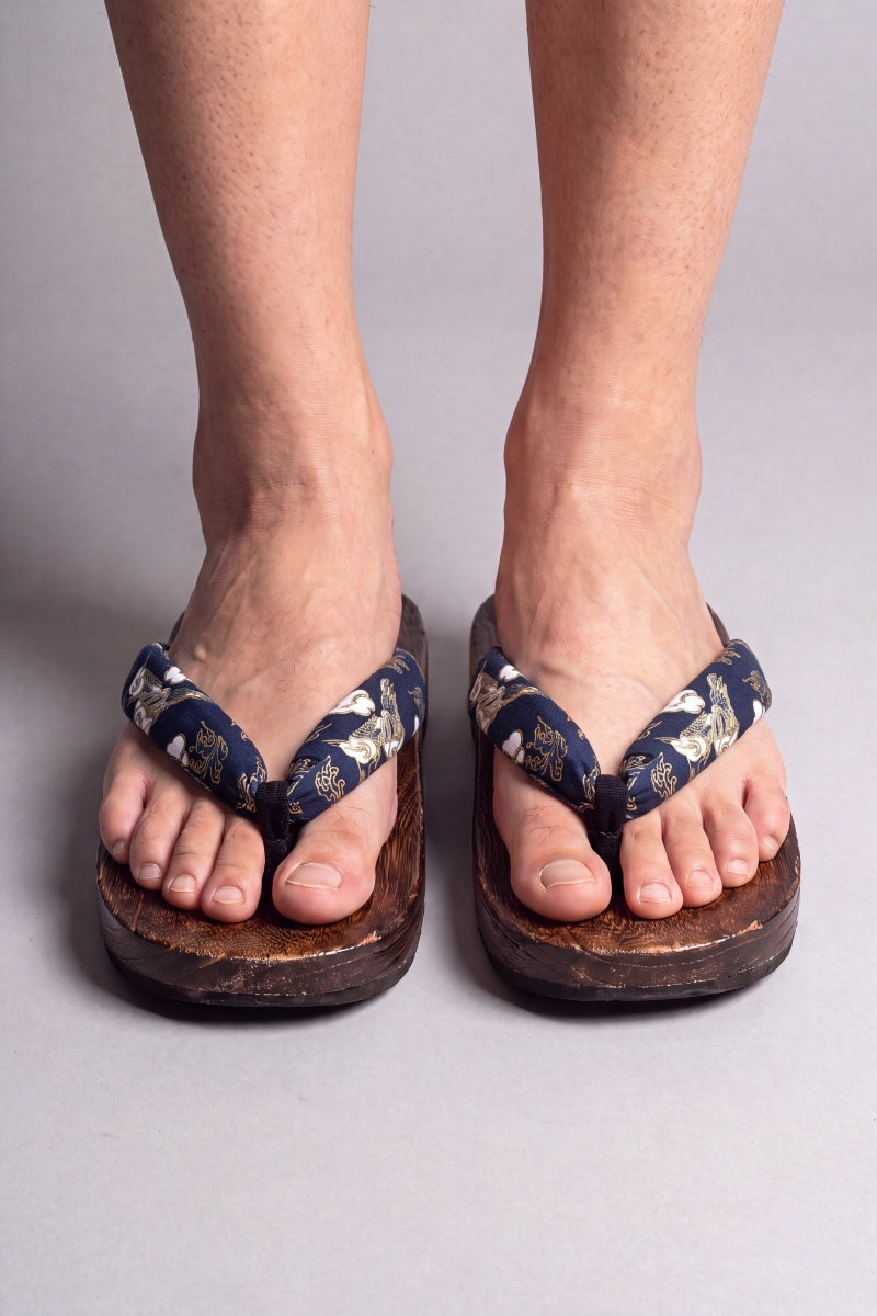 Chanclas japonesas color flores doradas sobre azul noche, calzadas descalzas de frente — suela de madera maciza marrón y tira acolchada entre los dedos