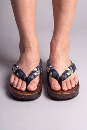 Chanclas japonesas color flores doradas sobre azul noche, calzadas descalzas de frente — suela de madera maciza marrón y tira acolchada entre los dedos