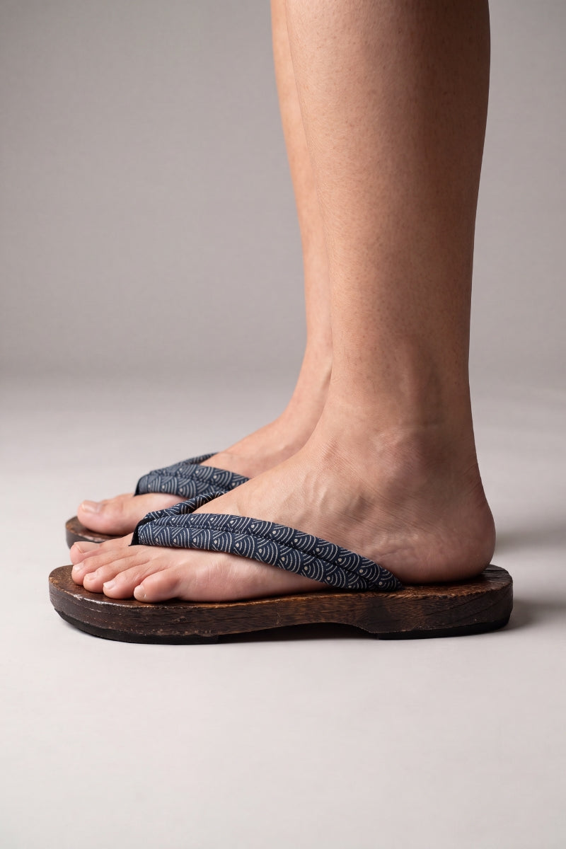 Chanclas japonesas vistas de perfil, calzadas descalzas — suela de madera plana y curvada, tira azul grisáceo con motivo de olas