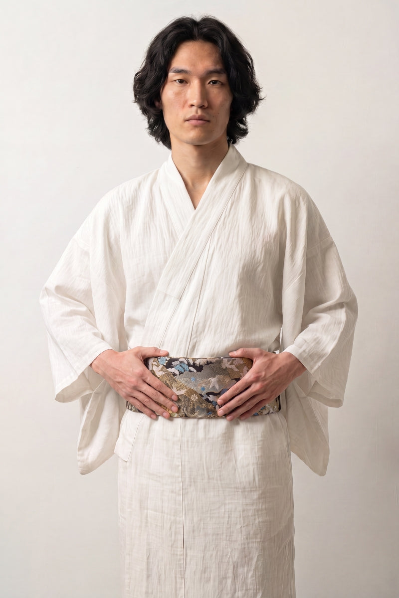 Cinturón obi 'Tachibana' ceñido a la cintura sobre un kimono blanco por un modelo masculino — brocado negro cubierto de flores plateadas y doradas en camafeo