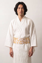 Cinturón obi 'Sanae' ceñido a la cintura sobre un kimono blanco por un modelo masculino — brocado crema y oro con volutas arremolinadas y flores blancas