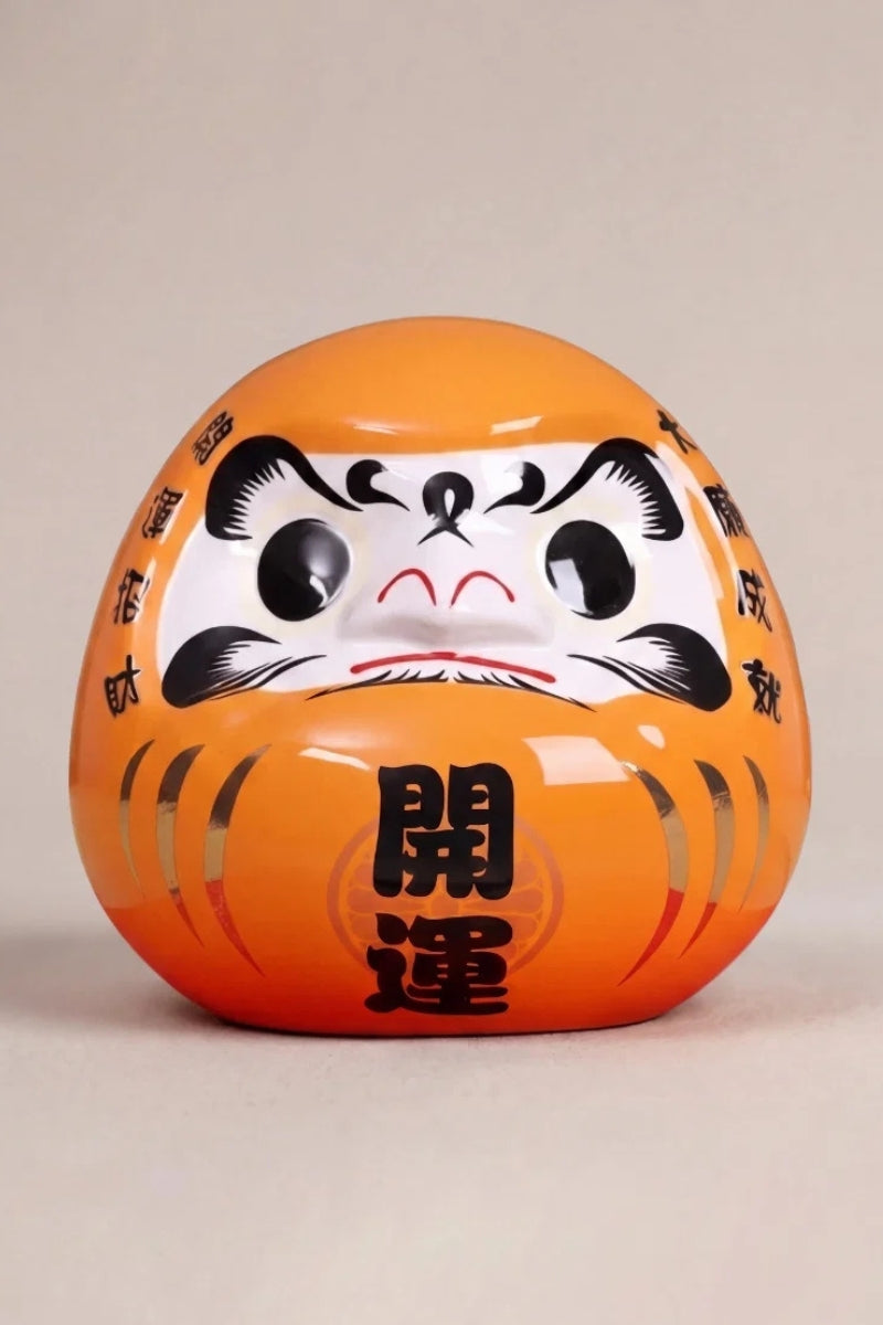 Daruma naranja imperial de cerámica sobre fondo beige — figura redonda de 11 cm con el kanji 皇, ojos en blanco para el rito del deseo