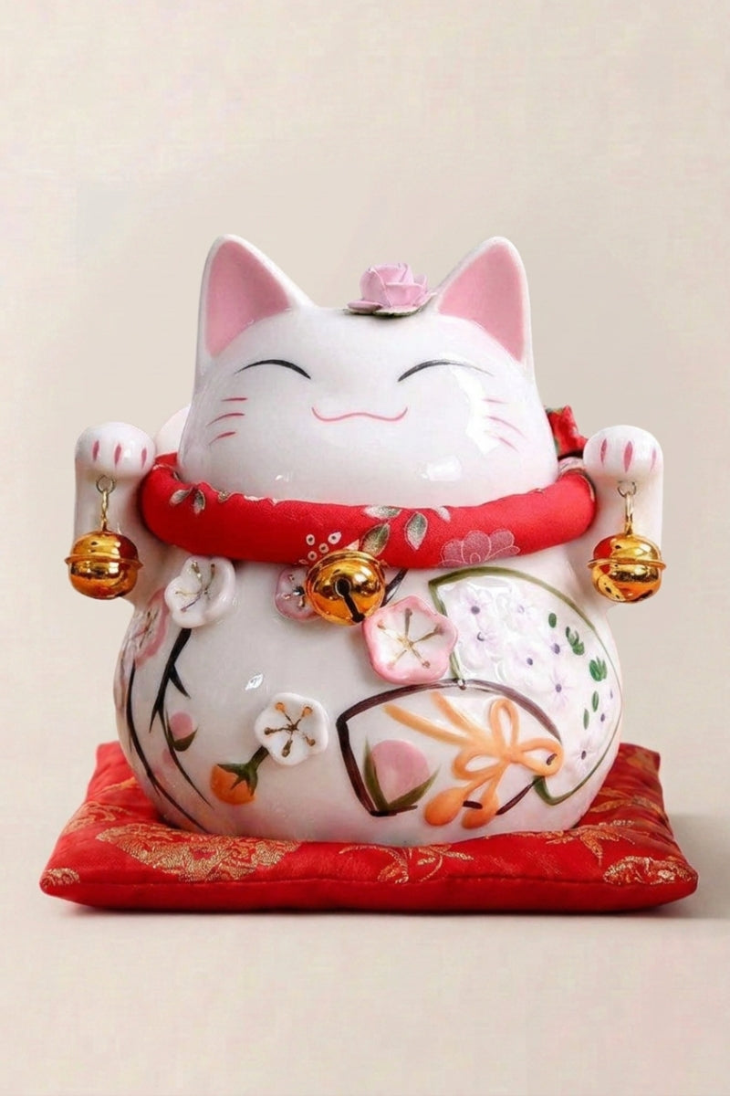 Hucha maneki neko 'Okami' de cerámica — gato blanco con los ojos cerrados, vientre con motivos florales y cascabeles dorados, sobre un cojín rojo