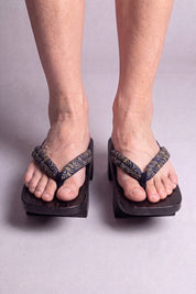 Geta japonés 'Tokoname' calzada con los pies descalzos, vista frontal — tira oscura con arabescos dorados sobre plataforma alzada de madera teñida de negro