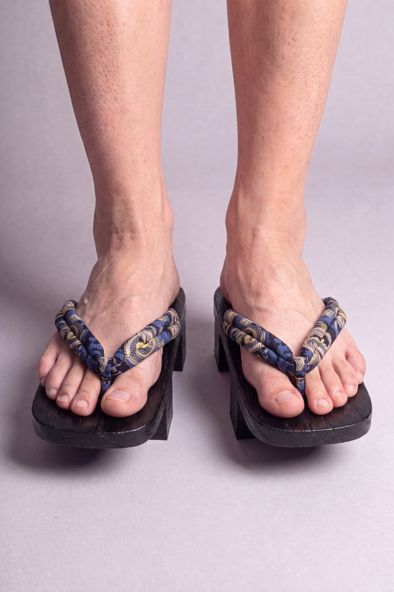 Geta japonés 'Nagoya' calzada con los pies descalzos, vista frontal — tira azul noche con volutas doradas sobre plataforma alzada de madera teñida de negro