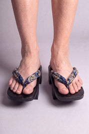 Geta japonés 'Nagoya' calzada con los pies descalzos, vista frontal — tira azul noche con volutas doradas sobre plataforma alzada de madera teñida de negro