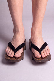 Geta japonés 'Iwakura' calzada con los pies descalzos, vista frontal — tira negra lisa sobre plataforma alzada de madera oscura