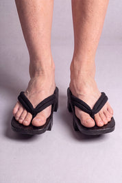 Geta japonés 'Tsushima' calzada con los pies descalzos, vista frontal — tira negra fina y flexible sobre plataforma alzada de madera teñida de negro