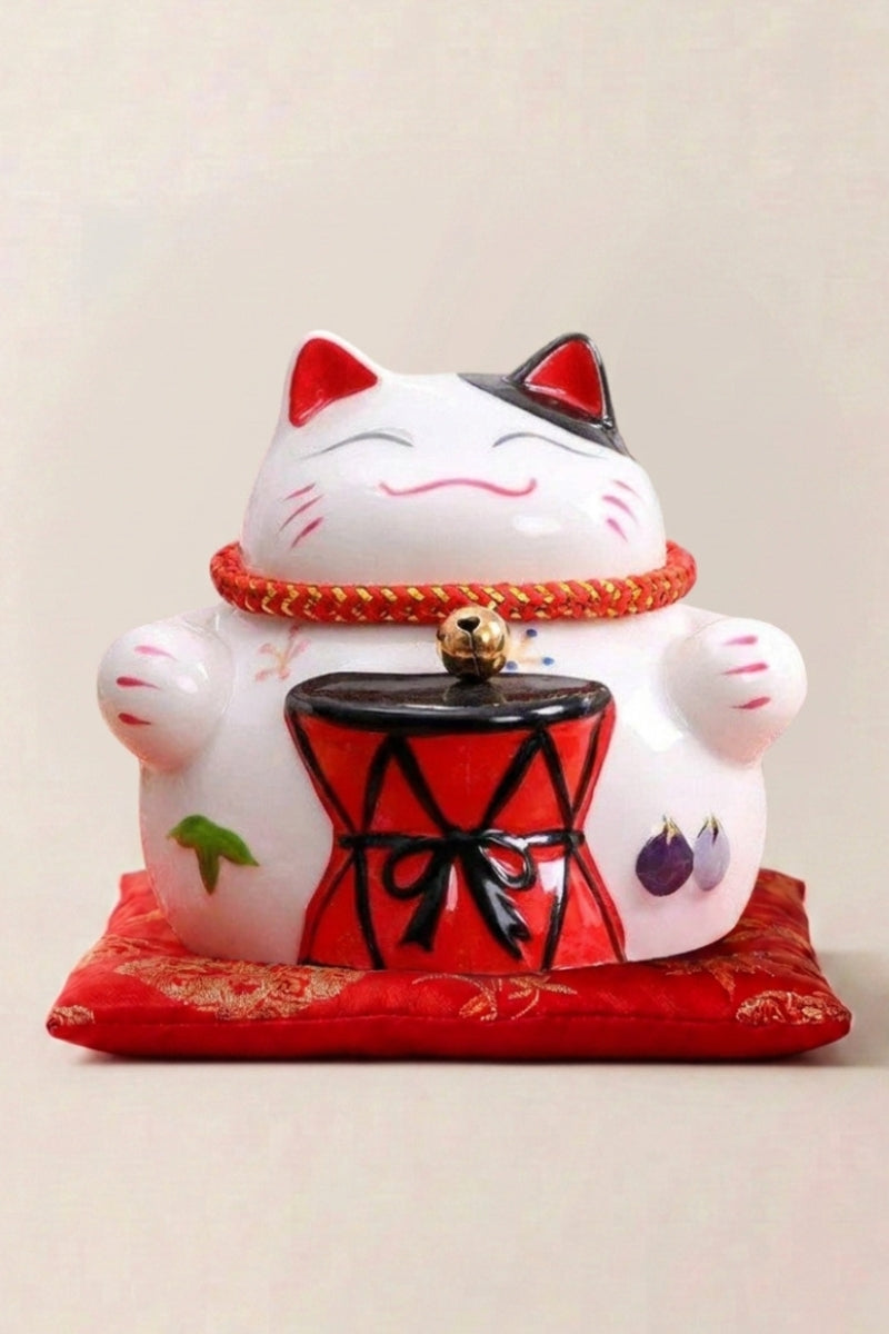 Hucha maneki neko 'Tambourine' de cerámica — gato blanco sosteniendo un tambor negro sobre soporte y collar con cascabel rojo, sobre un cojín rojo