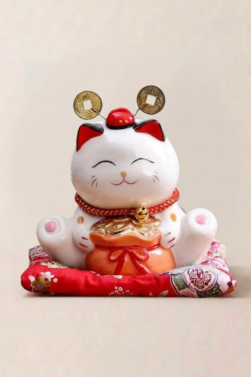 Hucha maneki neko 'Rich Cat' de cerámica — gato blanco rodeado de monedas doradas, orejas rojas y ambas patas alzadas, sobre un cojín rojo