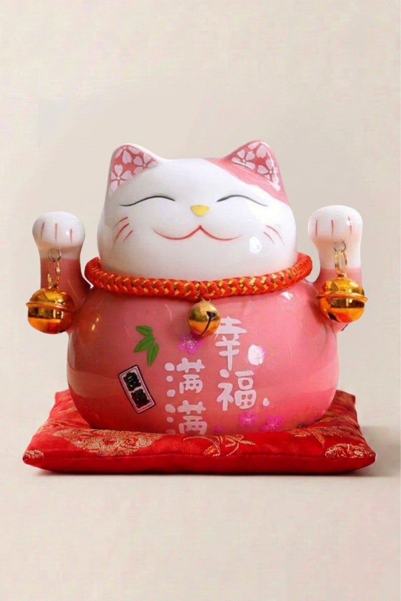 Hucha maneki neko 'Rose' de cerámica — gato rosa pálido con flores de cerezo y placa caligrafiada, ambas patas alzadas, sobre un cojín rojo