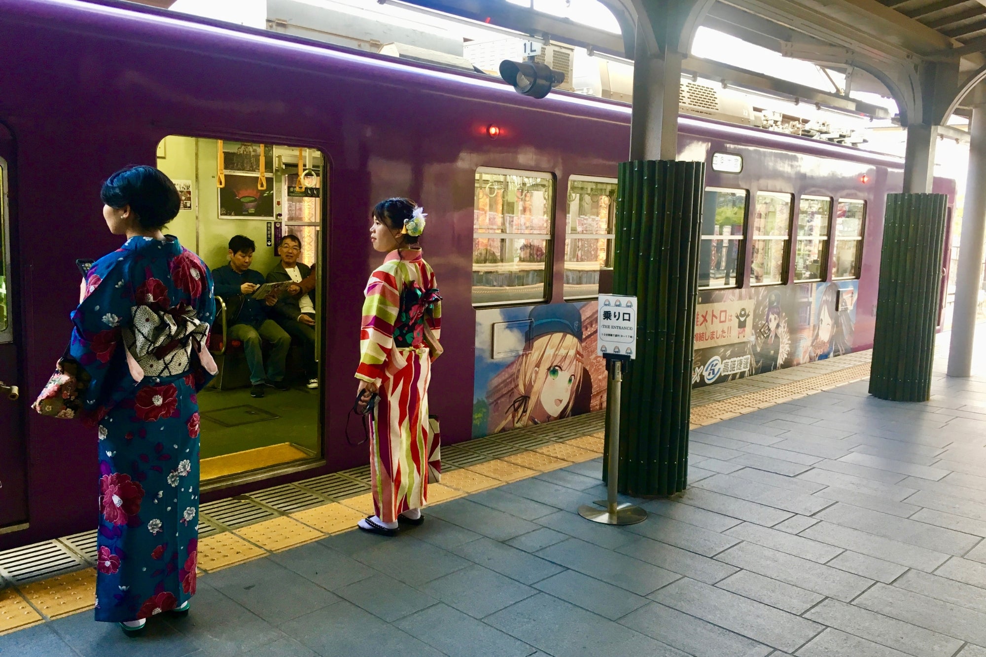 Dos mujeres vestidas con kimono esperan un tren violeta decorado con ilustraciones de anime en Kioto, entre tradición japonesa y cultura contemporánea.