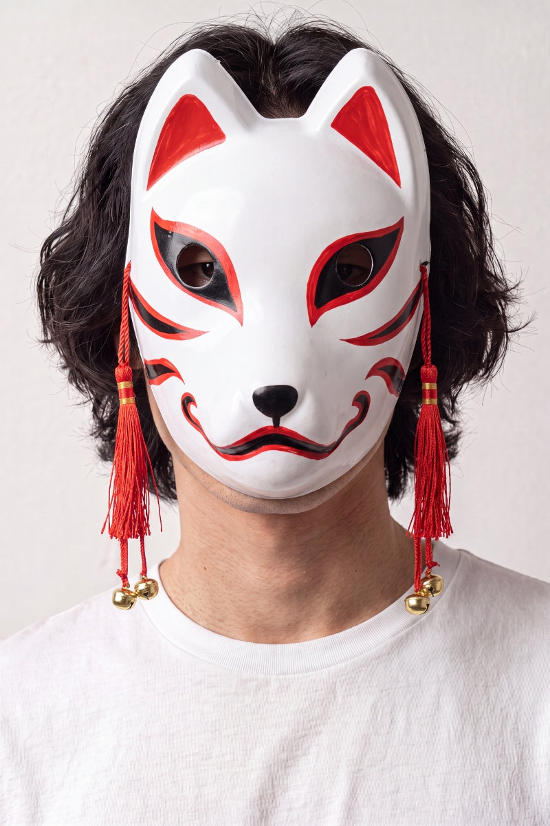 Máscara kitsune roja y blanca llevada por un modelo sobre fondo claro — zorro blanco con trazos rojos, borlas rojas y cascabeles dorados a los lados