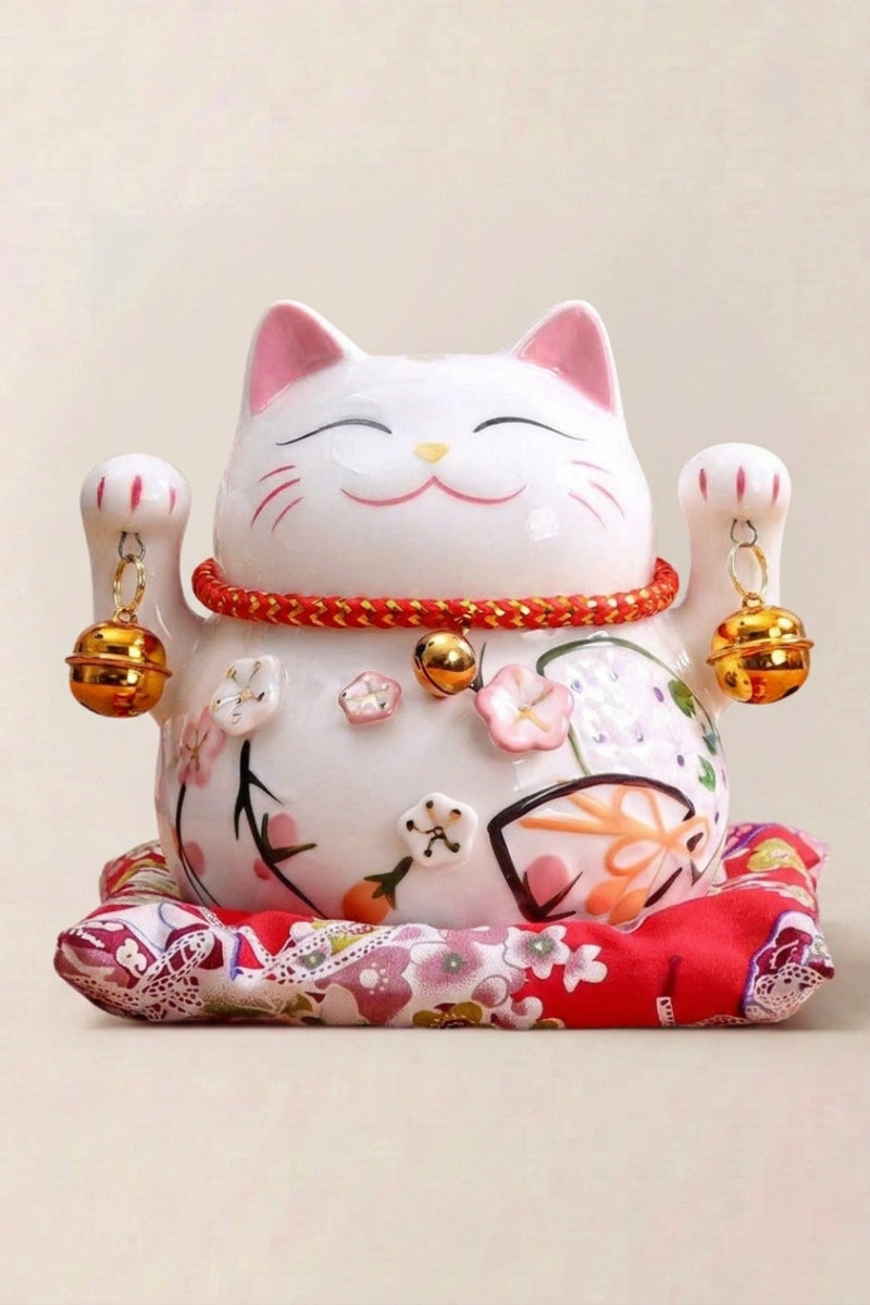 Hucha maneki neko 'Tinkerbell' de cerámica — gato blanco cubierto de cascabeles dorados y flores, ambas patas alzadas, sobre un cojín rojo
