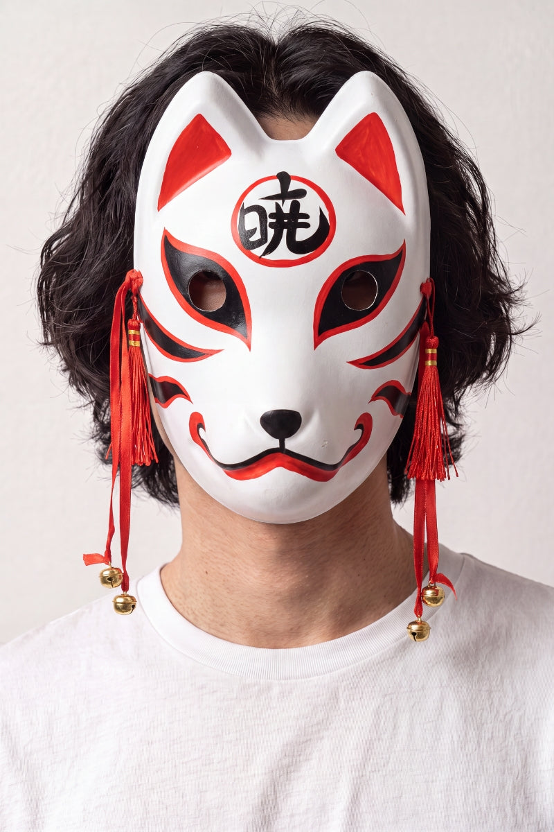 Máscara kitsune con kanji llevada por un modelo sobre fondo claro — zorro blanco con un kanji rojo en la frente, cordones y cascabeles dorados