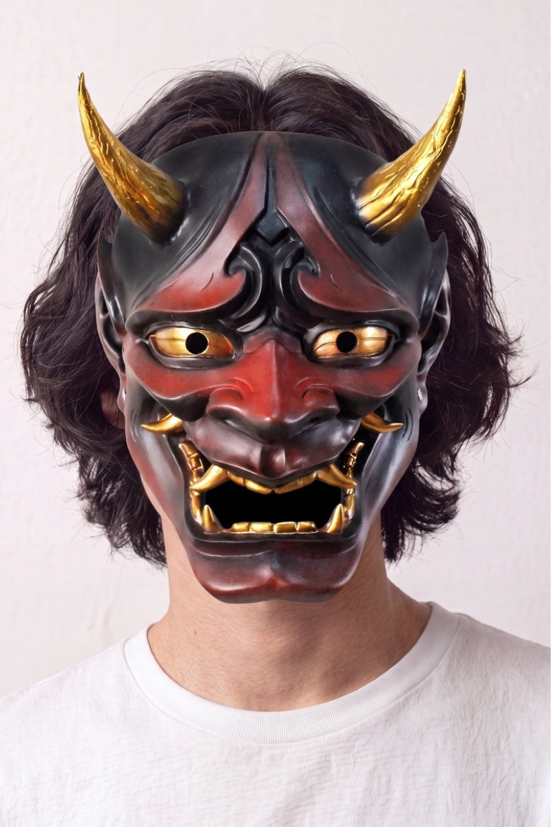 Máscara hannya roja llevada por un modelo sobre fondo claro — demonio rojo y negro con cuernos dorados, colmillos blancos y ojos ribeteados de oro