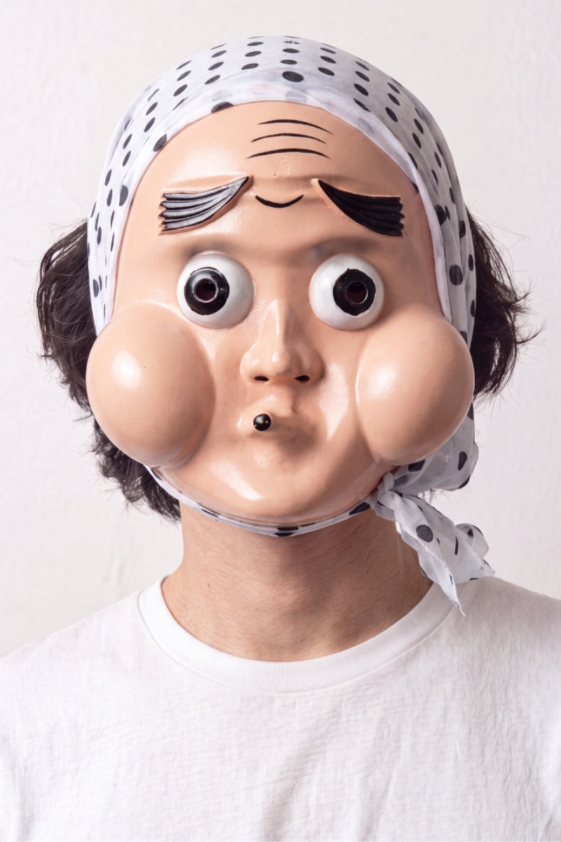 Máscara hyottoko llevada por un modelo sobre fondo claro — rostro cómico de mejillas redondas, boca fruncida de lado y pañuelo azul