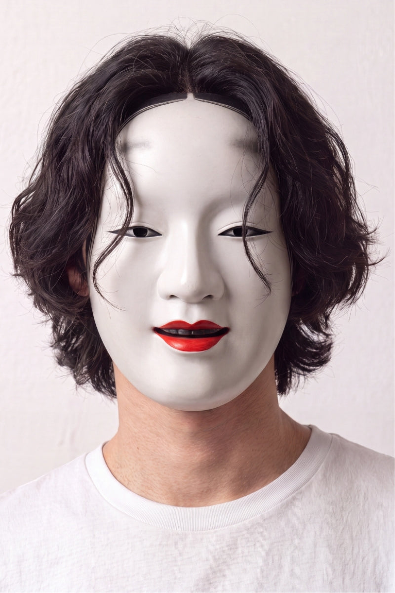 Máscara de nô llevada por un modelo sobre fondo claro — rostro femenino blanco lacado, labios rojos y cejas altas pintadas