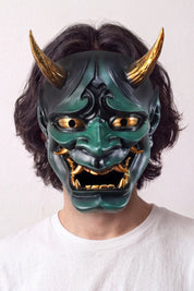 Máscara hannya verde llevada por un modelo sobre fondo claro — demonio verde con cuernos dorados, ojos saltones y mandíbula abierta con colmillos