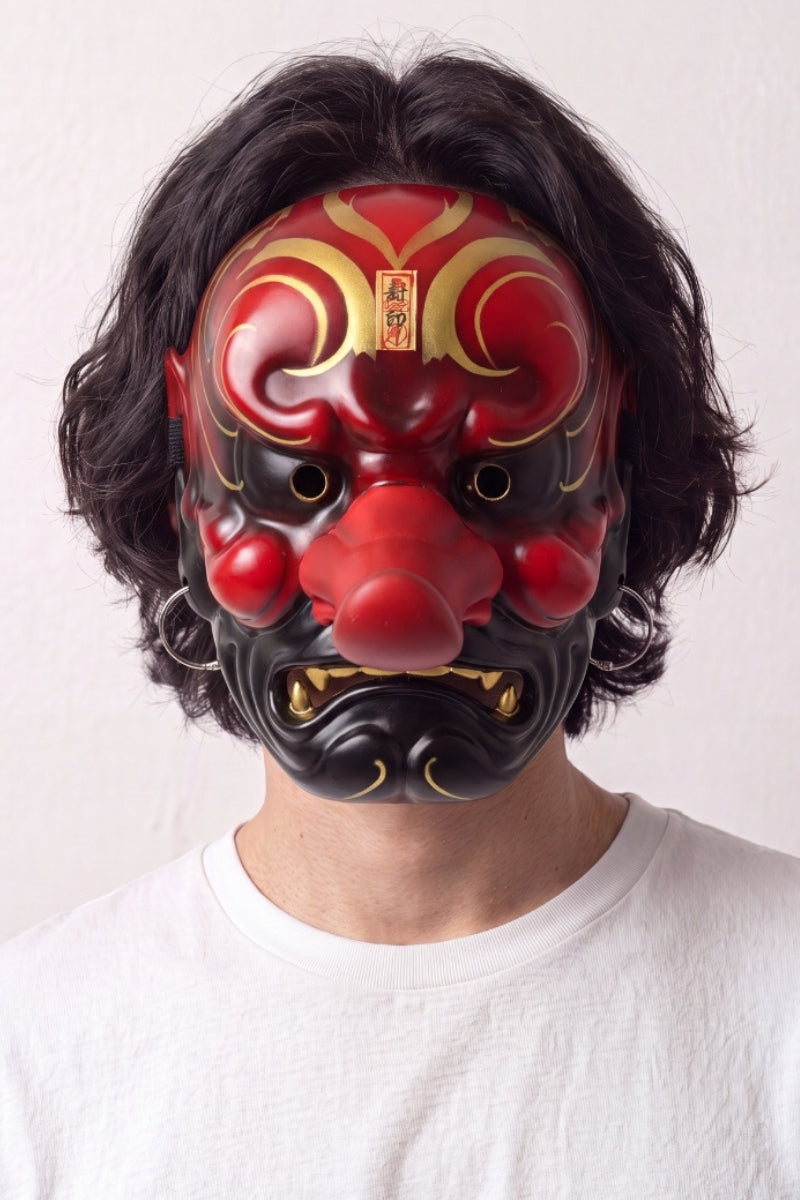 Máscara tengu llevada por un modelo sobre fondo claro — nariz larga y prominente, cejas en volutas doradas y colmillos blancos