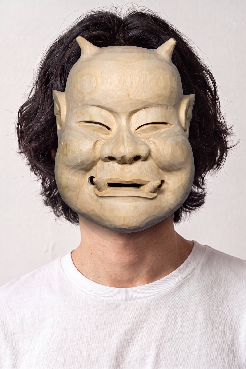 Máscara de nô beige llevada por un modelo sobre fondo claro — rostro anciano sonriente de ojos entornados, tono arena y modelado sobrio