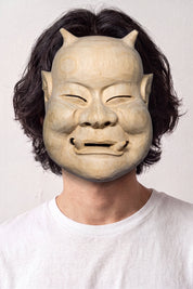 Máscara de nô beige llevada por un modelo sobre fondo claro — rostro anciano sonriente de ojos entornados, tono arena y modelado sobrio