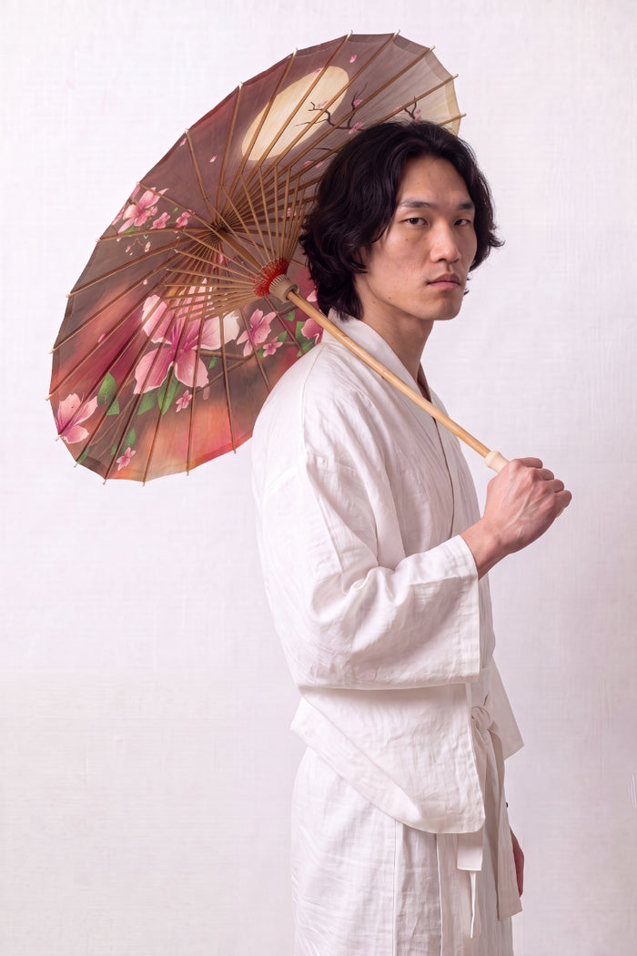 Sombrilla japonesa 'Chisana' abierta y sostenida por un modelo con kimono blanco — tela pequeña en degradado marrón y rosa con flores de cerezo