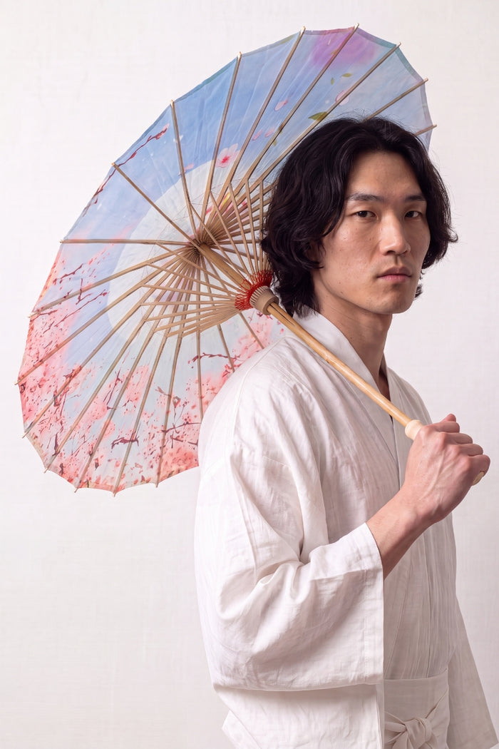 Sombrilla japonesa 'Kekkon' abierta y sostenida por un modelo con kimono blanco — tela azul claro y rosa con nubes estilizadas y flores
