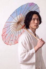 Sombrilla japonesa 'Kekkon' abierta y sostenida por un modelo con kimono blanco — tela azul claro y rosa con nubes estilizadas y flores