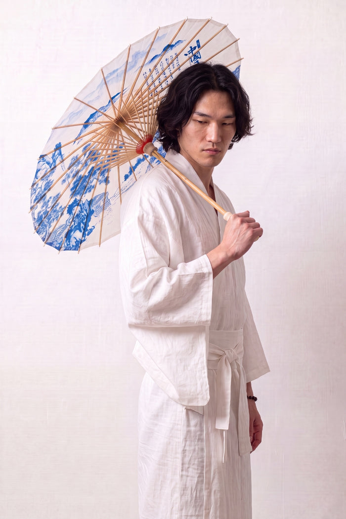 Sombrilla japonesa 'Kashika' abierta y sostenida por un modelo con kimono blanco — tela blanca con motivo de jardín en azul, follaje y pabellones