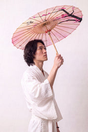 Sombrilla japonesa 'Sakura Fleuri' abierta y sostenida por un modelo con kimono blanco — tela rosa en degradado con rama oscura y flores de cerezo
