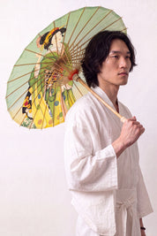 Sombrilla japonesa 'Kimiko' abierta y sostenida por un modelo con kimono blanco — tela verde oliva y amarilla con figuras de mujeres en kimono
