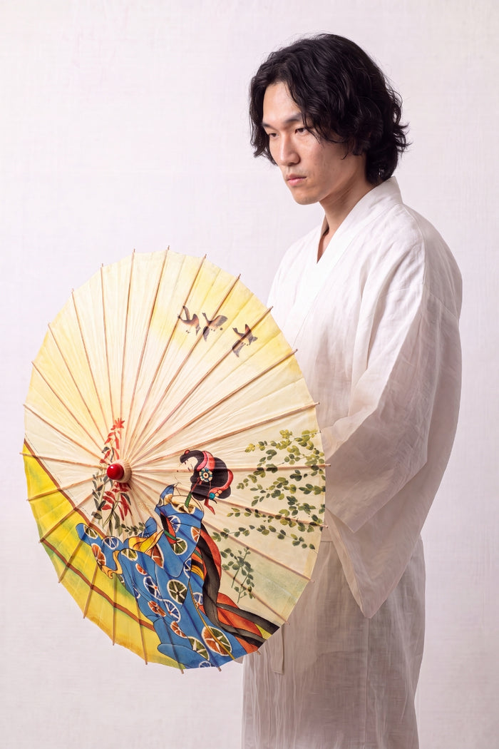 Sombrilla japonesa 'Shojino' abierta y sostenida por un modelo con kimono blanco — tela amarilla y azul con figura de maiko y ramas en flor