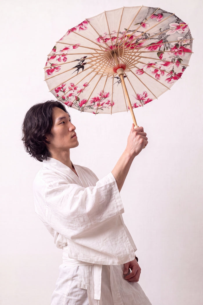 Sombrilla japonesa 'Nomikomu' abierta y sostenida por un modelo con kimono blanco — tela crema cubierta de flores de cerezo rosas