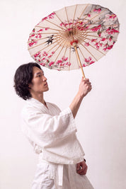 Sombrilla japonesa 'Nomikomu' abierta y sostenida por un modelo con kimono blanco — tela crema cubierta de flores de cerezo rosas
