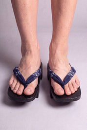 Geta japonés 'Okimari' calzada descalza, vista frontal — tira azul noche con motivo floral blanco sobre plataforma de madera teñida de negro