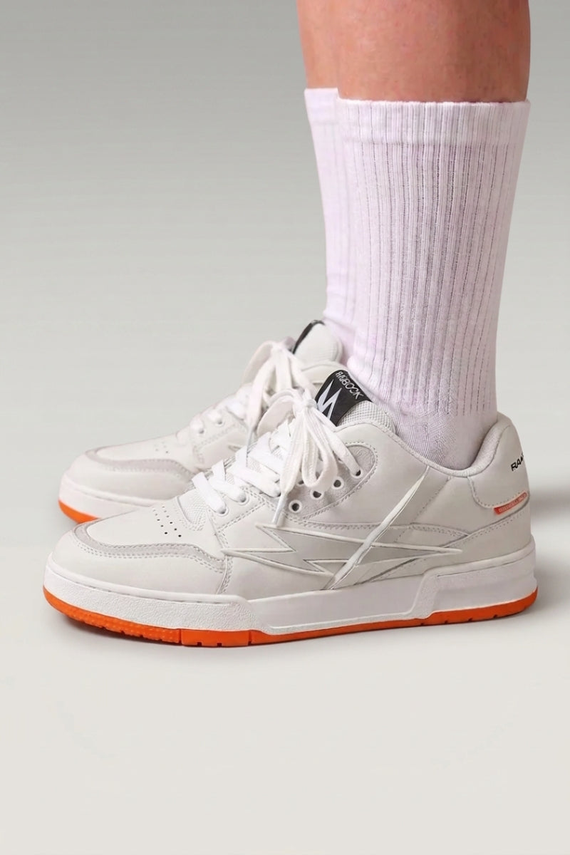 Zapatillas chunky 'Inazuma' en naranja — corte blanco y gris sobre una suela monocroma viva, con calcetines blancos sobre fondo de estudio gris