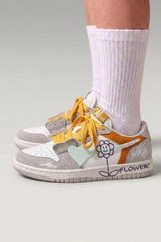 Zapatillas bajas 'Hanabi' en blanco — parches de color, flor cosida en el talón y cordones anchos, con calcetines blancos sobre fondo de estudio gris