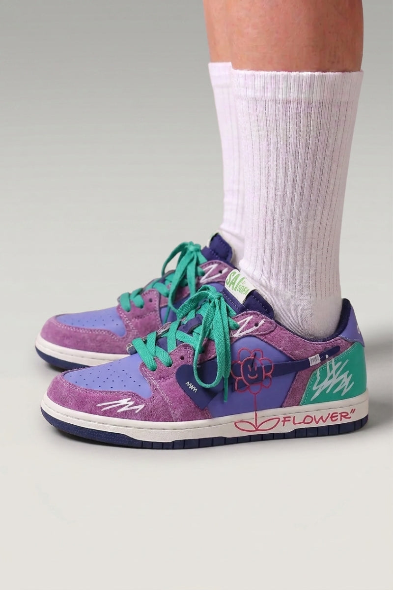 Zapatillas bajas 'Hanabi' en violeta — parches de color, flor cosida en el talón y cordones anchos, con calcetines blancos sobre fondo de estudio gris