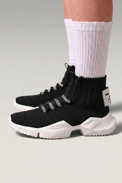 Zapatillas altas 'Kashiwa' en negro — punto alto tipo calcetín sin cordones y suela gruesa, con calcetines blancos sobre fondo de estudio gris