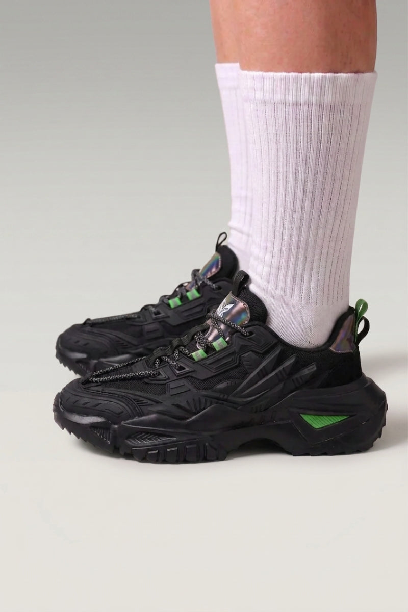 Zapatillas chunky 'Shinshiro' en negro — líneas nervadas en los laterales y suela moteada, con calcetines blancos sobre fondo de estudio gris