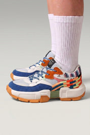 Zapatillas chunky 'Tsuruga' en blanco — paneles contrastados azul y naranja y suela de goma, con calcetines blancos sobre fondo de estudio gris
