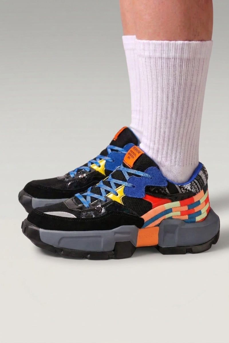 Zapatillas chunky 'Tsuruga' en negro — paneles contrastados azul y naranja y suela de goma, con calcetines blancos sobre fondo de estudio gris